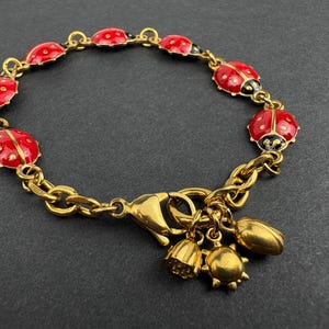 Könnte beinhalten: Ein goldfarbenes Kettenarmband mit mehreren rot-schwarzen Marienkäfer-Anhängern aus Emaille, die entlang der Glieder verteilt sind. Das Armband hat einen Karabinerverschluss und drei baumelnde goldfarbene Anhänger in Blüten-, Sonnen- und Blattform.
