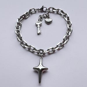 May include: Silver-toned chain bracelet with a star pendant. The bracelet features a series of oval links and includes charms of a key and a heart. The star pendant has a four-point design. The bracelet is displayed on a white background.