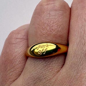 May include: A gold-toned metal ring featuring a smooth, oval-shaped face with the words "fuck off" engraved in a cursive script. The ring has a polished, reflective surface and a simple, solid band that encircles the finger.
