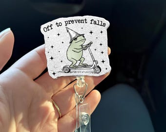 May include: A badge reel with a whimsical design featuring a frog riding a scooter and wearing a wizard hat. The text "Off to prevent falls" is printed above the frog. The badge reel has a clear plastic holder and a metal clip.