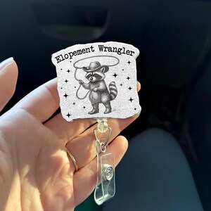 May include: A retractable badge reel featuring a cartoon raccoon in a cowboy hat, holding a lasso. The text "Elopement Wrangler" is above the raccoon. The reel has a clear plastic clip and a metal ring.