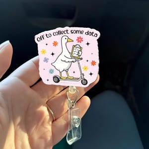 May include: A badge reel with a pink background featuring a cartoon goose riding a scooter and holding a clipboard. The text "Off to collect some data" is printed on the top. The design includes colorful flowers and stars.