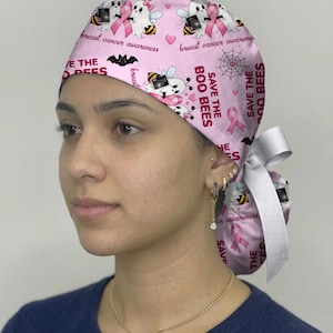 May include: A pink scrub cap featuring a repeating pattern of ghost bees, pink breast cancer awareness ribbons, black bats, and spider webs. The text reads "SAVE THE BOO BEES" and "breast cancer awareness". The cap has a white ribbon tie at the back.
