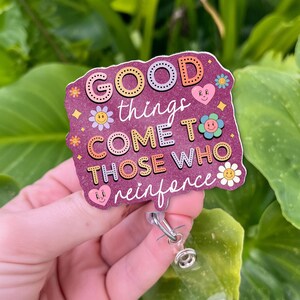 May include: A purple badge reel with the words "GOOD things COME TO THOSE WHO reinforce" in a colorful, dotted font. The badge has flower and heart graphics. The reel has a clear plastic strap and a metal clip.