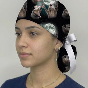 May include: A black scrub cap featuring a repeating pattern of opossums reaching toward a full moon against a starry night sky. The cap includes a white ribbon tie at the back to secure the fit, providing a functional head covering for medical professionals.