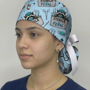 May include: A light blue scrub cap featuring a repeating pattern of raccoons wearing surgical masks and caps, medical instruments, and the text "STERILE but FERAL" printed on containers. The cap includes a white ribbon tie at the back.