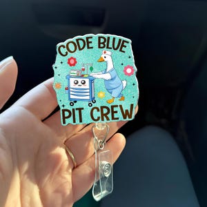 May include: A teal badge reel with the words "CODE BLUE PIT CREW" and a cartoon goose in a nurse's uniform. The goose is pushing a medical cart. The badge reel has a clear plastic strap and a metal clip.