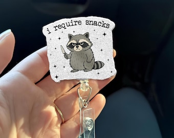 May include: A retractable badge reel with a cartoon raccoon holding a knife. The raccoon is gray and white with a black mask and a grumpy expression. The text "i require snacks" is above the raccoon.