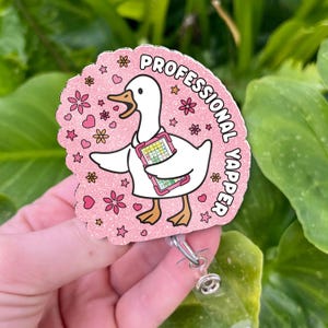 Puede incluir: Un portatarjetas rosa con un dibujo de un ganso sosteniendo una carpeta. El texto "PROFESSIONAL YAPPER" rodea al ganso. La insignia tiene un diseño floral y de estrellas.