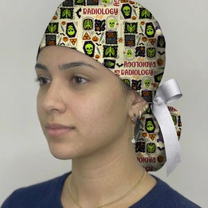 May include: A scrub cap featuring a cream background with a repeating pattern of skulls, skeletons, pumpkins, bats, and syringes. The text RADIOLOGY and SPOOKY NOTES is printed throughout. The cap includes a white ribbon tie at the back for adjustment.
