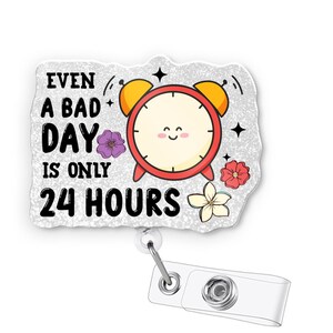 May include: A badge reel featuring the phrase "EVEN A BAD DAY IS ONLY 24 HOURS". The design includes a red alarm clock with a smiling face, yellow bells, and floral accents. The reel has a clear plastic clip.