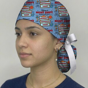 May include: A light blue scrub cap featuring a repeating pattern of clipboards with the text 'Welcome to NIGHT SHIFT WHERE EVERYDAY IS MONDAY YESTERDAY AND TODAY', coffee cups, and clocks, finished with a white ribbon tie at the back.