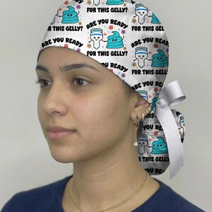 May include: A white scrub cap featuring a repeating pattern of cartoon gel pens and blue gel blobs with the text "ARE YOU READY FOR THIS GELLY?" in black, accented by small colorful flowers and hearts, finished with a white ribbon tie at the back.
