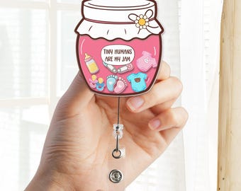 May include: A pink jar-shaped badge reel with a white lid and a flower accent. The jar contains baby-themed illustrations and the text "TINY HUMANS ARE MY JAM". The reel has a clear plastic clip and a retractable cord.