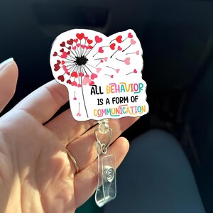 Op de afbeelding: Een witte badge reel met een paardenbloem ontwerp, met rode en roze hartvormige zaden. De tekst "ALL BEHAVIOR IS A FORM OF COMMUNICATION" wordt in kleurrijke letters weergegeven. De reel heeft een doorzichtige plastic houder en een metalen clip.
