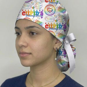 May include: A white satin scrub cap with a colorful "Cupid's Delivery Service" design. The cap features hearts, rainbows, and the text "Wrapped with Love" and "Hand Delivered With Care". A white satin bow is tied at the back.