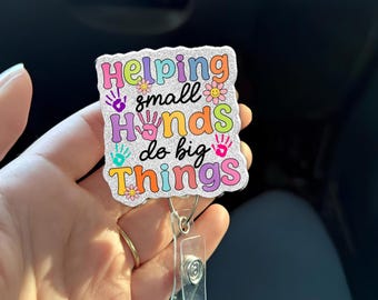 Może przedstawiać: Błyszcząca odznaka z napisem "Helping small hands do big things" w kolorowych literach. Odznaka ma przezroczysty plastikowy klips i chowany sznurek.