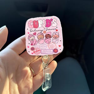 May include: A pink badge reel with the text "Cupid's Delivery Service" and illustrations of babies in pink swaddling clothes. The badge reel has a clear plastic holder and a metal clip.