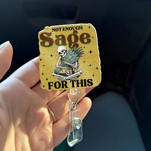 Puede incluir: Un portatarjetas con fondo dorado y las palabras "NOT ENOUGH Sage FOR THIS". La insignia presenta un esqueleto sosteniendo un manojo de salvia. El portatarjetas tiene una correa de plástico transparente y un clip de metal.