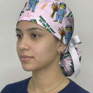 May include: A light pink scrub cap featuring a repeating pattern of green frogs wearing blue medical scrubs, holding bouquets of flowers, and adhesive bandages. The text SORRY FOR WHAT MY FACE SAID is printed in green, with red EKG heart rate lines.