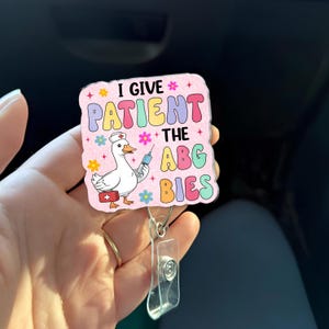 May include: A colorful badge reel featuring a cartoon goose in a nurse's hat holding a syringe. The text on the badge reads "I GIVE PATIENT THE ABG BIES" in various colors. The reel has a clear plastic clip and a metal retractable cord.