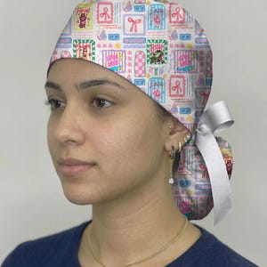 May include: A patterned surgical scrub cap featuring a repeating design of framed illustrations, bows, and perfume bottles in pastel colors. The cap is tied with a white ribbon. The overall theme is whimsical and feminine.