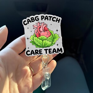 Puede incluir: Un portatarjetas con el texto "CABG PATCH CARE TEAM". La insignia presenta un corazón rosa sobre una cama de lechuga verde. El portatarjetas tiene un soporte de plástico transparente y un clip plateado.