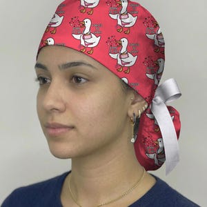 May include: A pink scrub cap featuring a repeating pattern of white ducks wearing stethoscopes and carrying clipboards. The text "BORN TO YAP" and "FORCED TO CHART" is printed near each duck. The cap includes a white ribbon tie at the back.