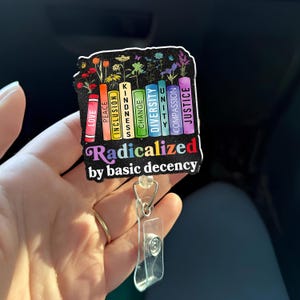 May include: A badge reel with a black background featuring colorful books labeled with words like "Love," "Kindness," and "Justice." The text "Radicalized by basic decency" is also displayed. The reel has a clear plastic holder and a metal clip.