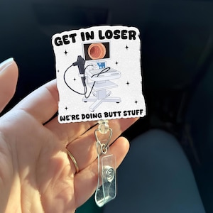 May include: A white badge reel with the text "GET IN LOSER" and "WE'RE DOING BUTT STUFF". The badge features a medical illustration of a colonoscopy machine. The reel has a clear plastic holder and a silver-colored clip.