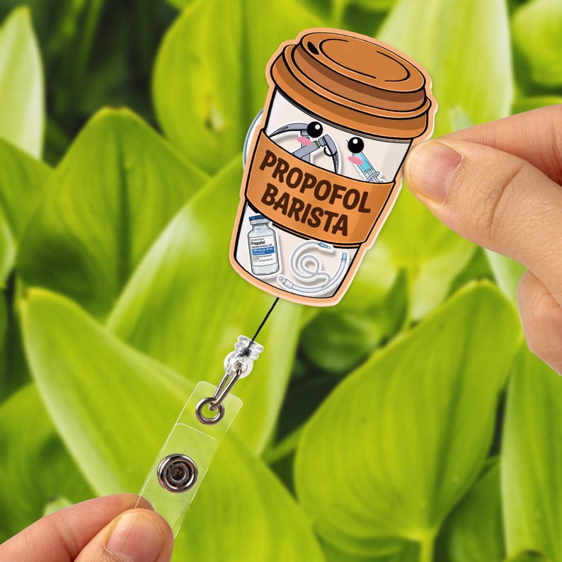 May include: A badge reel featuring a cartoon coffee cup design with the text "PROPOFOL BARISTA." The cup includes medical-themed illustrations such as a syringe and a vial. The reel has a clear plastic strap and a metal clip.
