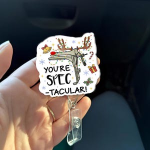 May include: A white badge reel with a medical speculum design, decorated with Christmas lights, antlers, and the text "You're Spec-tacular!". Includes a clear plastic badge holder and a metal clip.