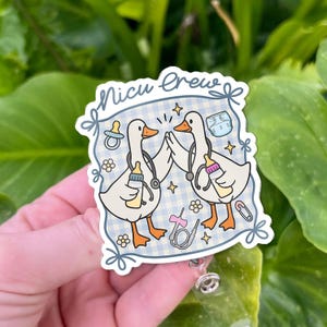 May include: A badge reel with a design of two cartoon geese wearing stethoscopes, holding baby bottles, and high-fiving. The badge reel has the text "Nicu Crew" and is decorated with baby-related icons on a blue and white checkered background.