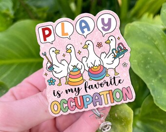 Könnte beinhalten: Ein rosa Ausweis-Rollhalter mit dem Wort „PLAY“ in Sprechblasen über vier Cartoon-Enten. Die Enten halten Spielzeug und der Text „is my favorite OCCUPATION“ steht darunter. Die Rolle hat einen transparenten Kunststoffriemen und einen Metallclip.
