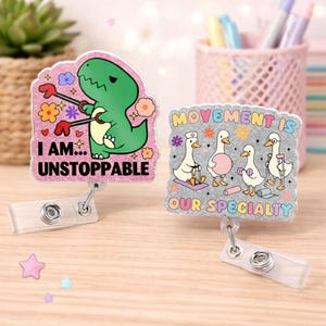 Puede incluir: Dos carretes de insignia brillantes. Uno presenta un dinosaurio verde con el texto "I AM... UNSTOPPABLE". El otro tiene cuatro patos de dibujos animados con el texto "MOVEMENT IS OUR SPECIALTY". Ambos tienen un clip de plástico transparente.