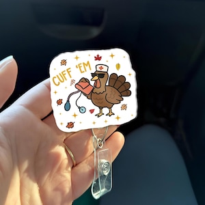 May include: A square badge reel featuring a cartoon turkey wearing sunglasses and a nurse's hat, holding a stethoscope and a medical bag. The text "CUFF 'EM" is printed on the top left. Autumn leaves and stars decorate the edges.
