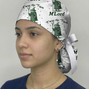 May include: A white scrub cap featuring a repeating dark green graphic print of a frog wearing a wizard hat and robe, holding a staff. The text "M'Lord" and "T'was the 'Tism" is printed in dark green. The cap includes a white ribbon tie at the back.