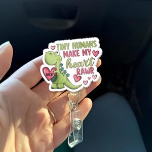 May include: A badge reel with a cartoon dinosaur holding a heart that says "NICU". The badge reel has the text "TINY HUMANS MAKE MY HEART RAWR" in green and pink. The badge reel has a clear plastic clip and a metal reel.