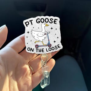 Puede incluir: Un portatarjetas blanco con un dibujo de un ganso en un patinete. El texto "PT GOOSE ON THE LOOSE" está impreso en la tarjeta. El portatarjetas tiene una correa de plástico transparente y un clip metálico.