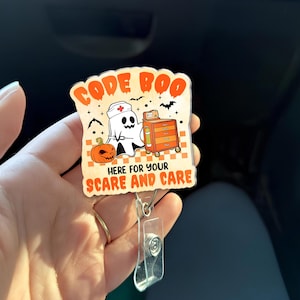 Puede incluir: Un portatarjetas con temática de Halloween con las palabras "CODE BOO" en letras naranjas. El diseño presenta un fantasma con gorro de enfermera, una calabaza y el texto "HERE FOR YOUR SCARE AND CARE". El portatarjetas tiene un clip de plástico transparente.