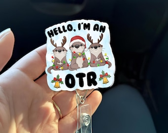 May include: A white badge reel featuring three cartoon otters with antlers and Christmas lights. The otters are wearing Santa hats. The text "HELLO, I'M AN OTR" is displayed on the badge.