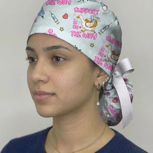 May include: A light blue scrub cap featuring a repeating pattern of white geese wearing medical gear, syringes, hearts, stars, and the text "SUPPORT IS ON THE WAY" in pink. The cap includes a white ribbon tie at the back to secure the fit.
