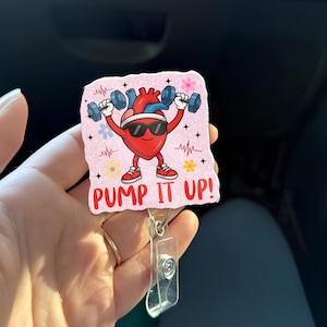 May include: A pink badge reel with a cartoon heart wearing sunglasses and lifting weights. The heart is red with blue accents and red sneakers. The text "PUMP IT UP!" is at the bottom. The reel has a clear plastic clip and a metal ring.