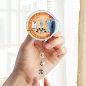 May include: A round, eye-shaped badge reel featuring a detailed anatomical illustration of the eye. The design includes an eye chart, eyeglasses, and a bottle of eye drops. The reel has a clear plastic strap and a metal snap.