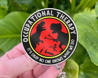 Könnte beinhalten: Ein runder Anstecker mit dem Text "OCCUPATIONAL THERAPY" und "IT'S OKAY NO ONE KNOWS WHAT WE DO". Der Anstecker zeigt zwei schwarze Dinosaurier-Silhouetten auf rotem Hintergrund, mit Werkzeugen. Er ist an einer ausziehbaren Rolle befestigt.