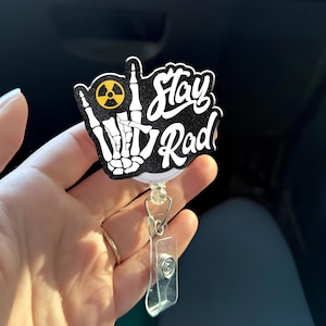 May include: A black and white badge reel with the words "Stay Rad" in white script. The design includes a skeleton hand giving the "hang loose" sign and a yellow radiation symbol. The reel has a clear plastic clip and a metal retractable cord.