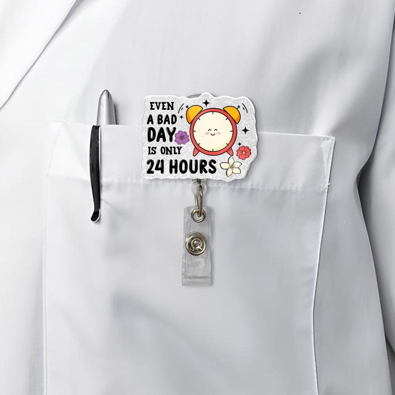 May include: A white badge reel featuring a cartoon alarm clock design and the text "Even a bad day is only 24 hours." The badge reel has a clear plastic clip and is attached to a white shirt pocket with a pen.