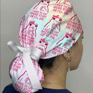 May include: A scrub cap featuring a pastel tie-dye background with a repeating pattern of pink frogs wearing bows. Each frog is accompanied by the text, I HAVE TO SAY WEIRD STUFF OR I'LL DIE. The cap includes a white ribbon tie at the back.