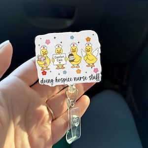 May include: A white badge reel with a design featuring four yellow cartoon ducks dressed as nurses. The ducks are holding flowers, a sign that says "Comfort Care", a bottle of medicine, and have angel wings. The text "doing hospice nurse stuff" is below the ducks.