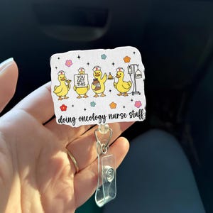 May include: A white badge reel with yellow cartoon ducks wearing nurse hats. The ducks are holding medical items and a sign that reads "YOU GOT THIS." The text "doing oncology nurse stuff" is at the bottom. The badge reel has a clear plastic holder and a metal clip.